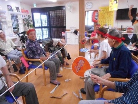 真ん中にひもを張った両側に、手にうちわを持って椅子に座った入居者が並ぶ共用スペース