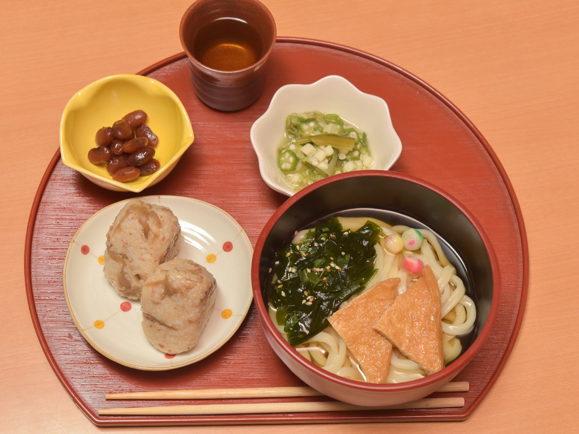 赤いお盆の上に食事がのっている。和雨とおあげの入ったうどんと、おにぎりが2個、オクラの和え物、豆がある。箸も用意してある。