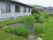 左にグレーの壁の平屋の建物があり、その右側に青い花が植えられている花壇