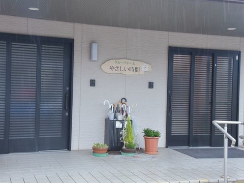 グレーの壁面に看板がかかっており、その両側に黒い横格子の3枚引き戸がある玄関