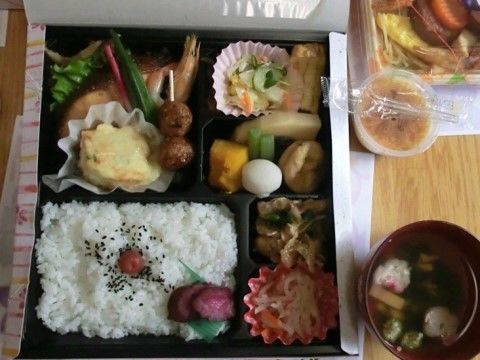 四角い黒い弁当箱にご飯や様々なおかずが盛られ、吸い物とデザートの付いた食事例