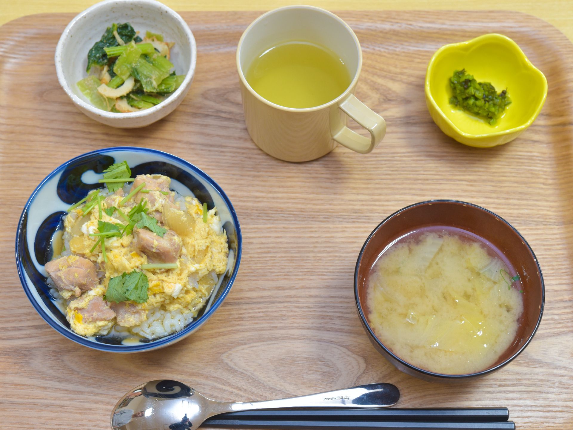 木製のトレーの上に箸とスプーンがセットされ、丼物、味噌汁、小鉢、漬物の4品と、マグカップのお茶が乗せられています。