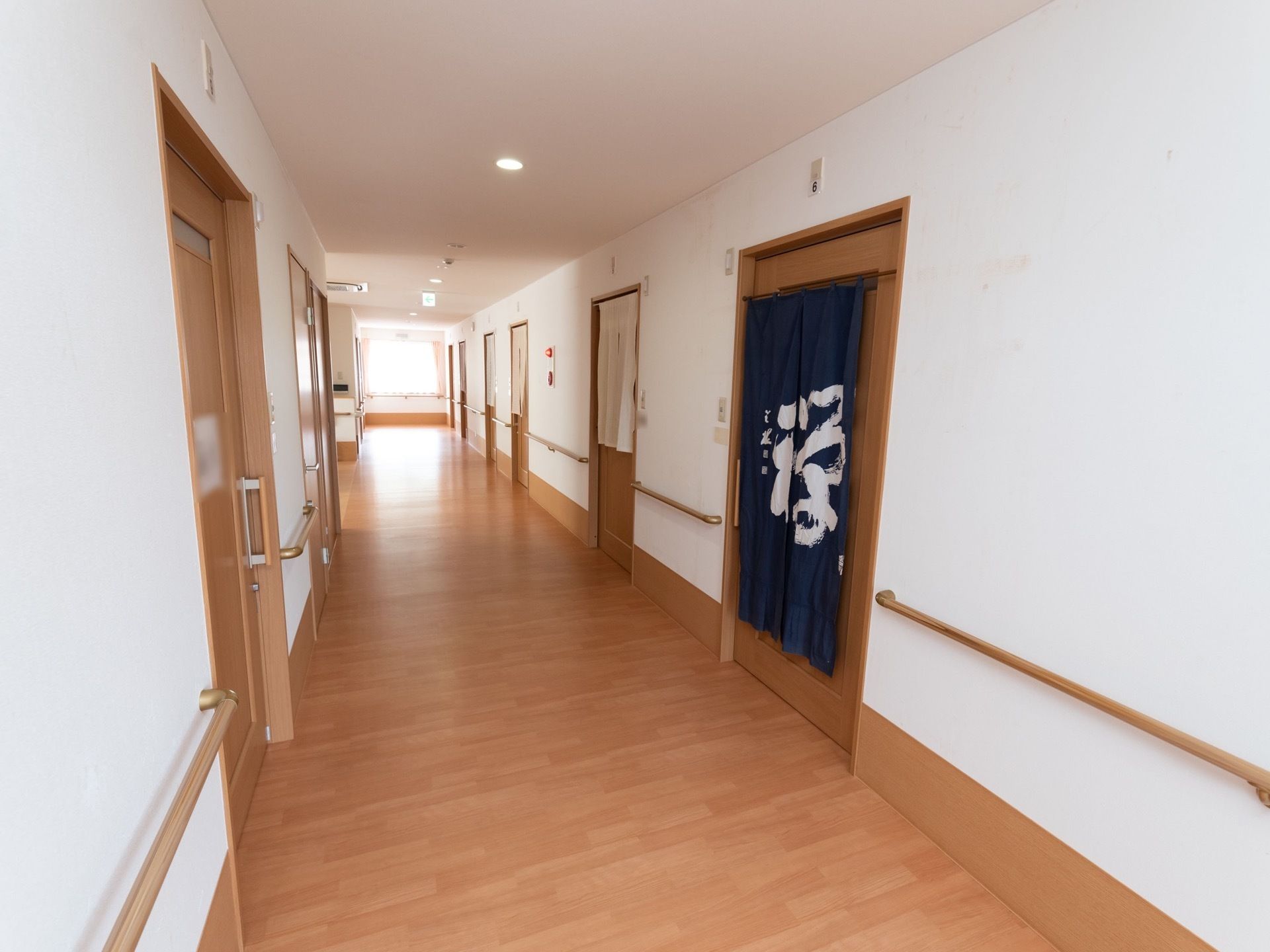 通路の床や居室のドアは暖かみのある色合い。通路の幅は広く、両側に手すりが設置されている。ドアの一部には暖簾が掛けられている。