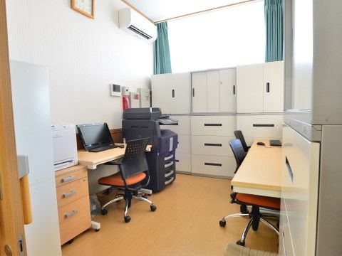 机にはノートパソコンが置かれ、椅子が置かれている。奥にある窓には緑のカーテンが掛かり、その手前に大きな書庫と黒の複合機が置かれている。