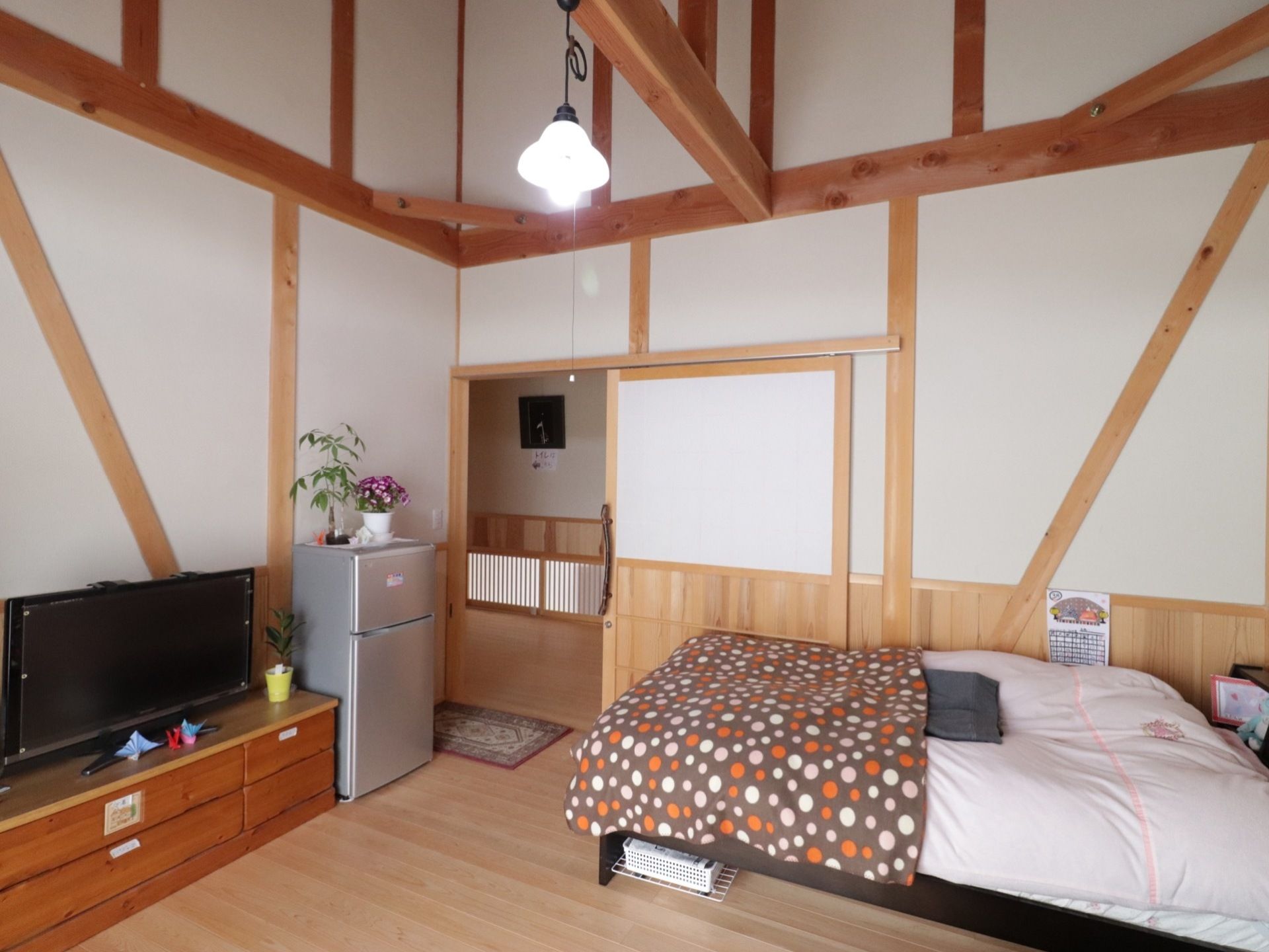 吹き抜けの天井の居室である。ベッドやテレビ、冷蔵庫が置かれている。照明は素朴なデザインの白熱灯である。