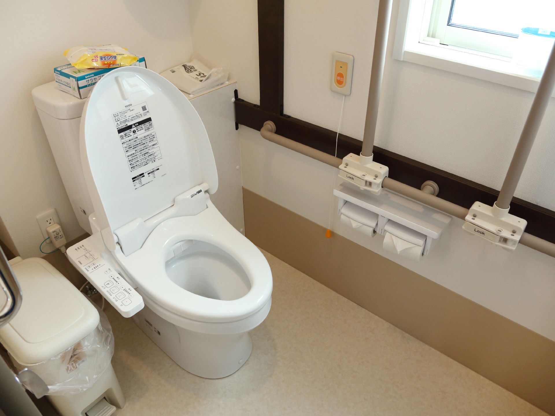 自然光の入り込む明るいトイレスペース。大きめのペダル式汚物入れを置いている。壁には可動式の手すりをつけている。