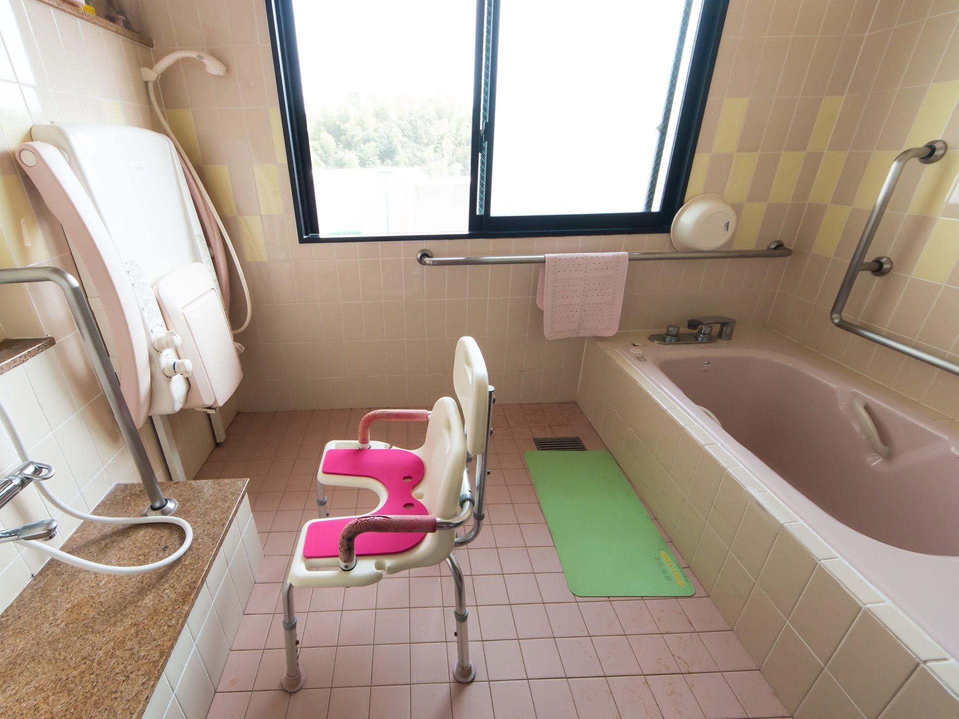 ピンクを基調としたタイルに囲まれた浴室がある。大きな窓が付いている。壁には手すりがあり、入浴介護用の機材が置かれている。