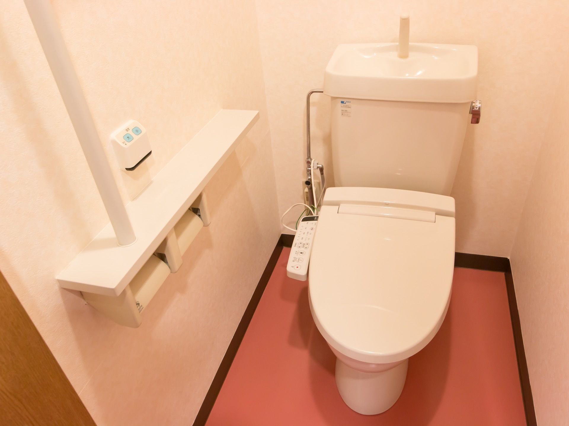 白い温水洗浄便座器が完備されたトイレである。壁には、呼び出しブザーと手すり付きのカウンターが設置されている。