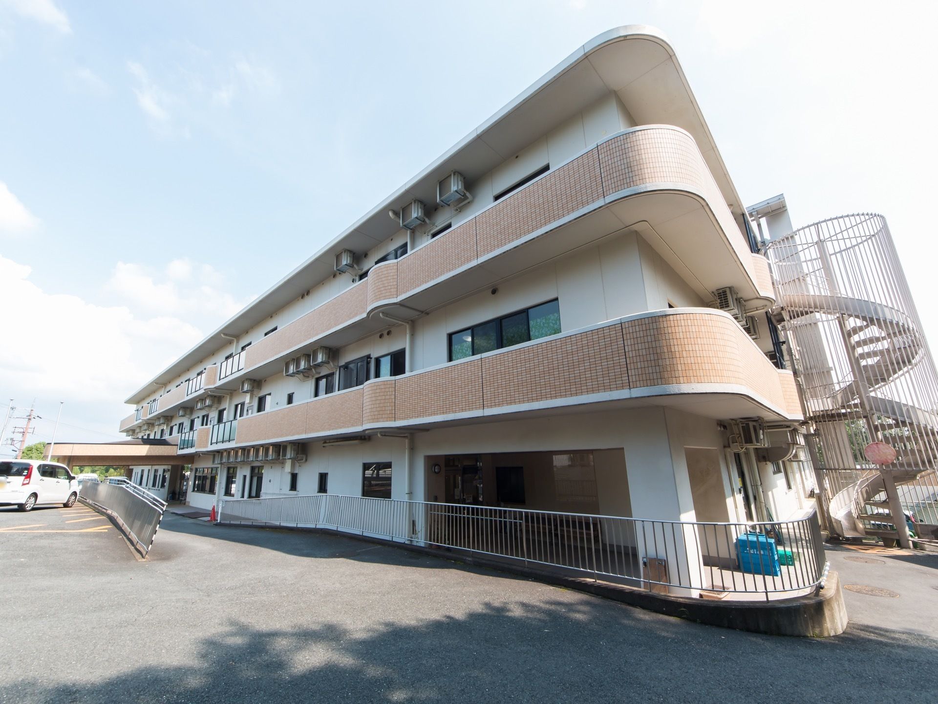 3階建ての施設外観である。入口へとゆるやかな勾配が付けられている。らせん状の非常階段が設置されている。