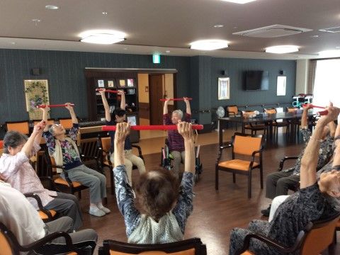 椅子に座って手に持った赤いものを引っ張りながら両腕を上げる運動に取り組む高齢者