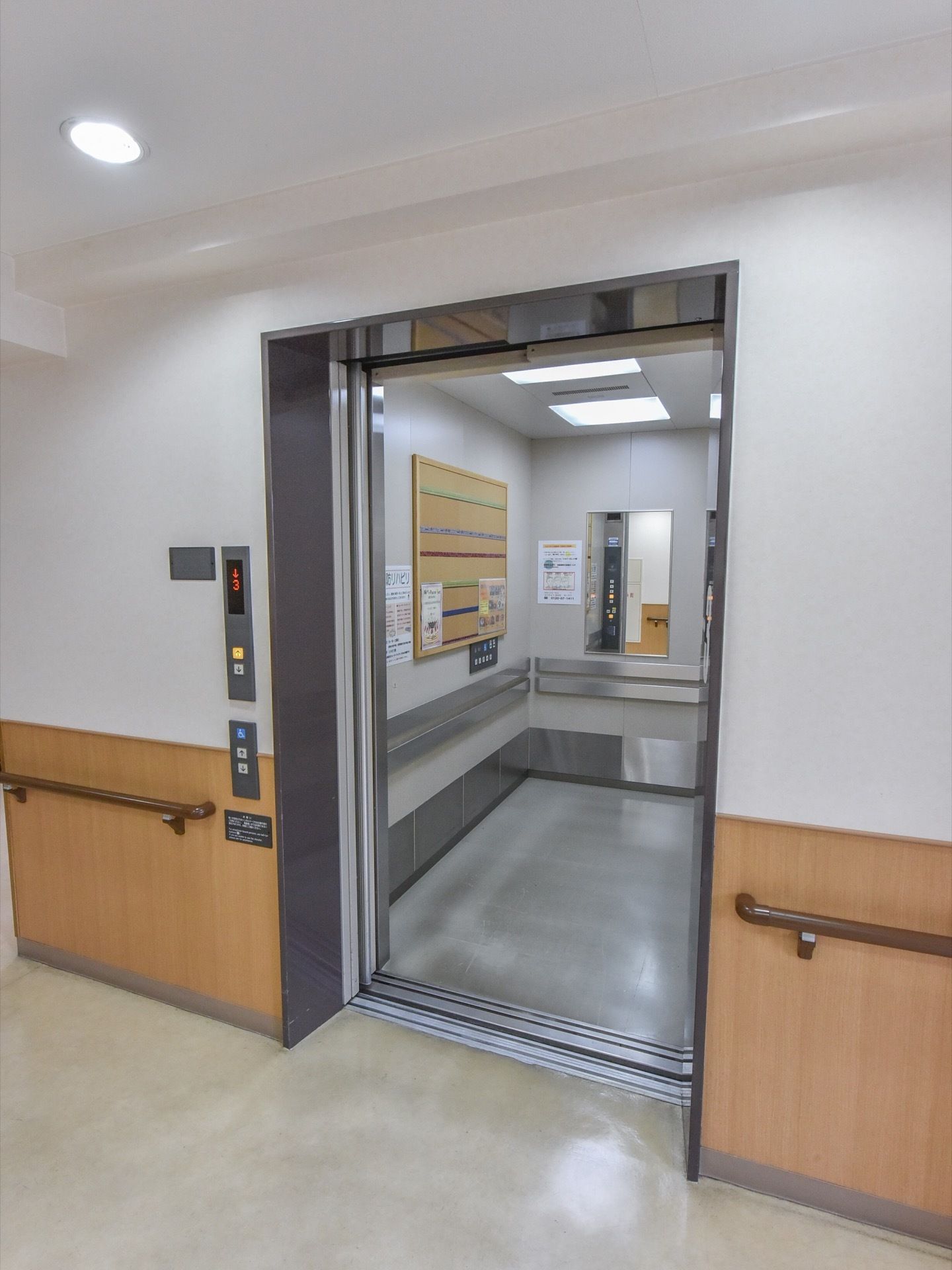 エレベーターは扉が開いた状態となっている。室内は電気がついていて奥行きがあり、掲示板や鏡が取り付けられている。