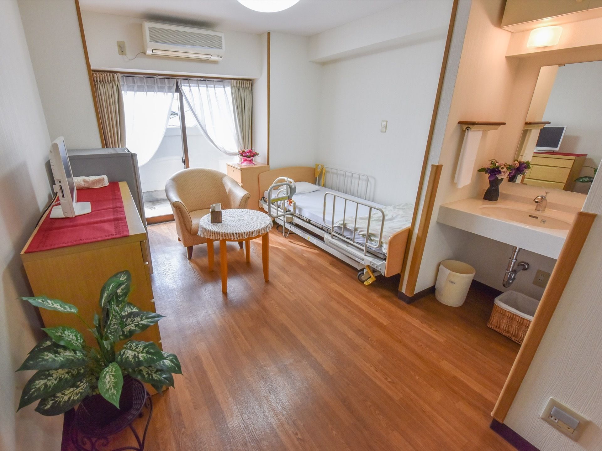 居室には丸型のテーブル、イス、ベッド、タンスといった家具が置いてあり、手前には観葉植物も飾られている。