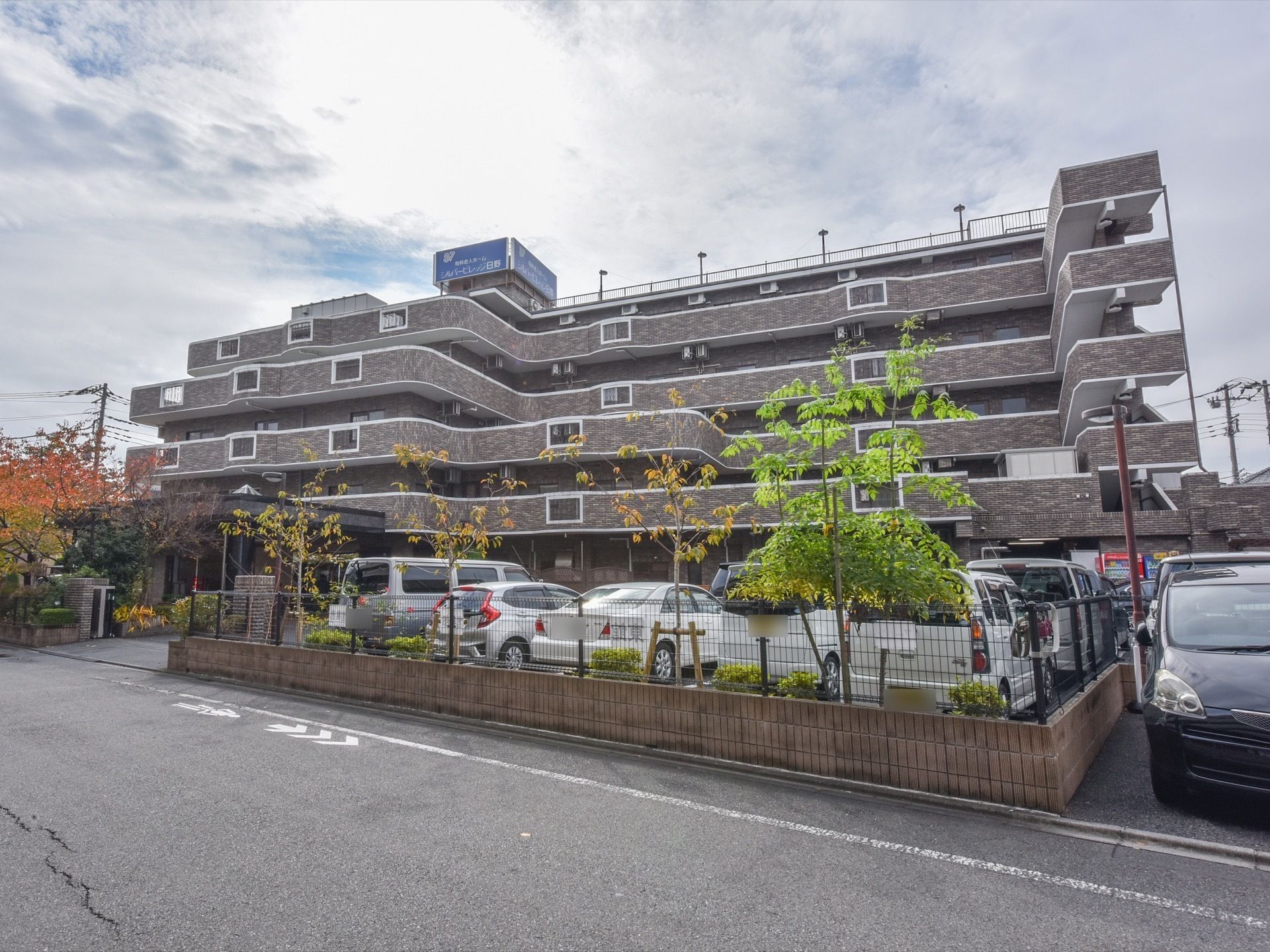 施設の外部分には広い駐車場があり、多くの車が柵の向こう側に停まっている。所々に色とりどりの木々が生えている。