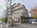 施設の入口部分から外観を見た形となっており、5階建ての建物になっている。地上部分には道路があり、車が停まっている。