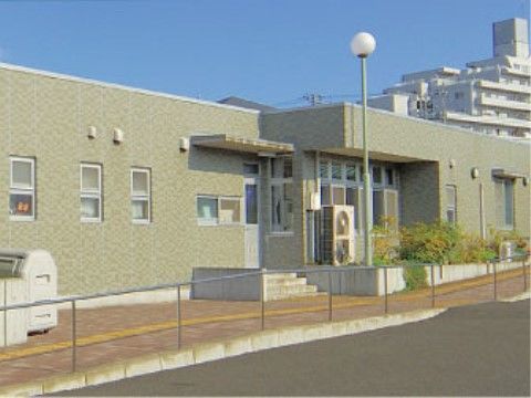 手前にオレンジ色のレンガ張り風の通路がある、薄い緑の壁の平屋の建物