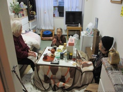 奥に窓があり、その手前にこたつが出されて3人の高齢者が話している居室