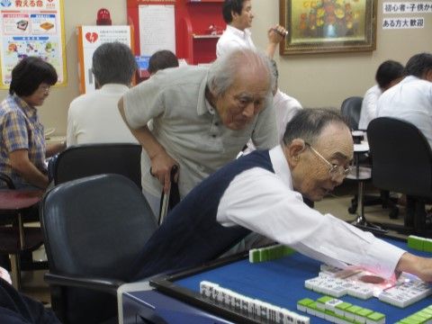 青い雀卓でマージャンをしている男性と、それを後ろからのぞき込んでいる男性