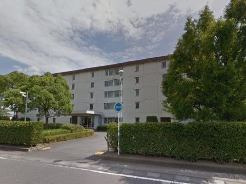 白い壁の５階建てのビルのような建物である。雲がかかった青空の下に大きな樹木が生い茂っている。敷地の入り口には街灯が立てられている。