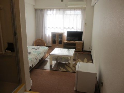 レースカーテンが掛けられた窓際に黒い薄型テレビとチェストが置かれている。ベッドと四角い座卓があり、ワンドアの冷蔵庫が設置されている。