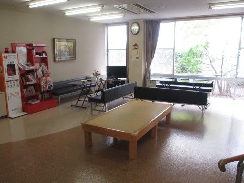 大きな窓からの光と天井に設置された蛍光灯に照らされた明るい部屋である。壁にAEDと本棚があり、部屋の中央にテーブルと椅子が置いてある。