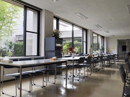 大きな窓から木漏れ日の入る明るい部屋に、長テーブルとパイプ椅子が並べられている。テーブルの上にティッシュの箱があり、花が飾られている。