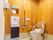 木目調の素材の壁に囲まれた広いトイレがある。小さな洗面台と鏡が設置されている。便座には背もたれがあり、壁には手すりがついている。