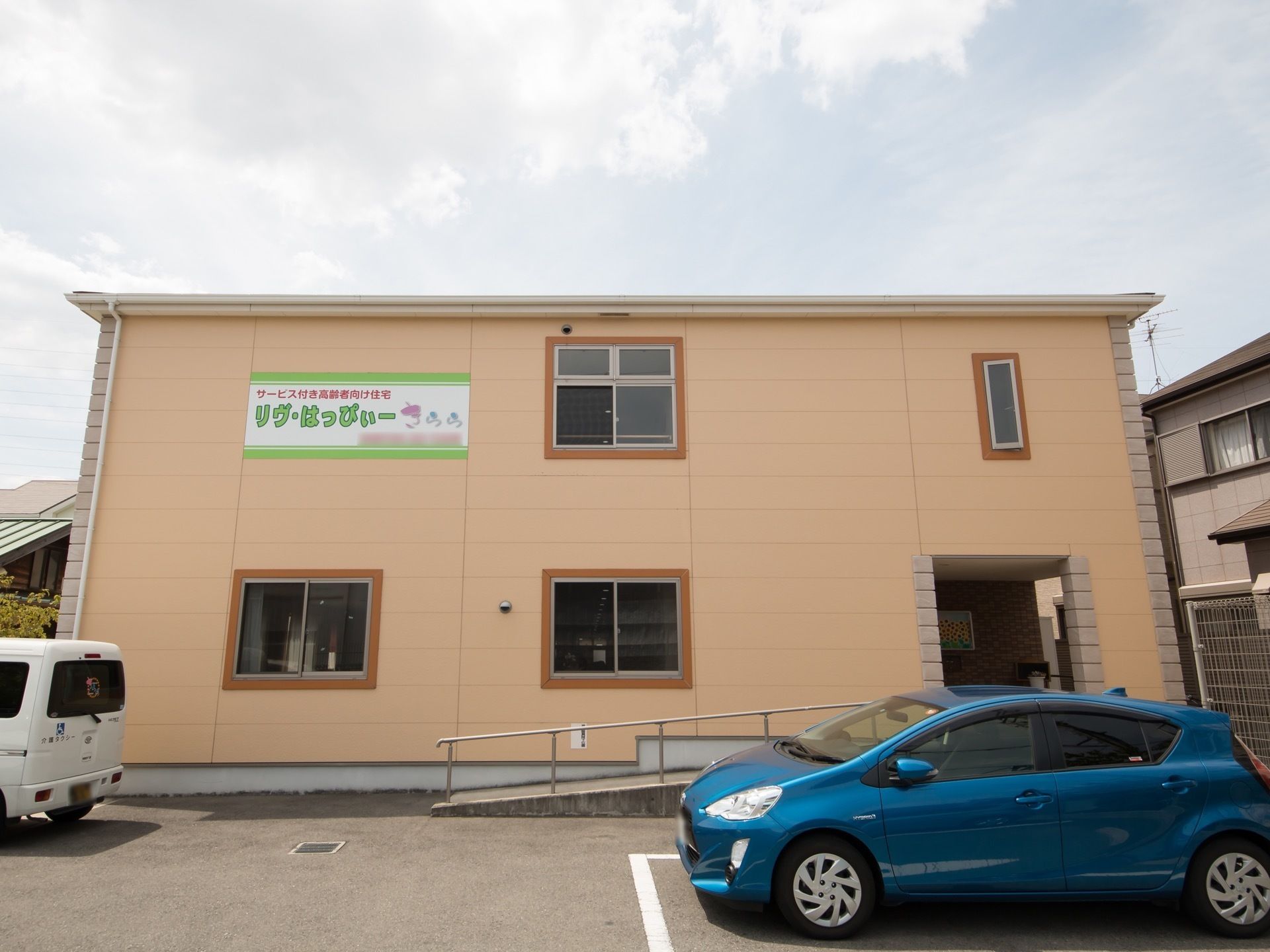 ２階建てのパステルカラーの建物があり、２階の外壁に施設名が表示されている。駐車場に青い車が停められている。