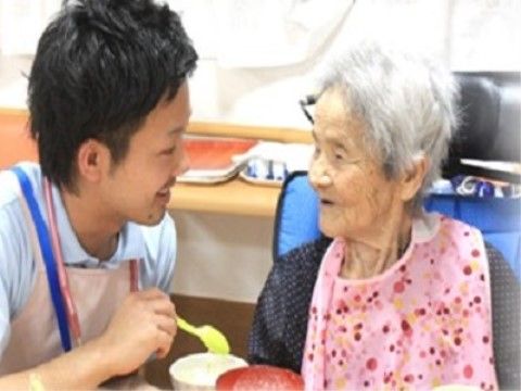 青い椅子に座って食事をしている白髪の高齢女性と、その左でスプーンを持って介助しているスタッフ