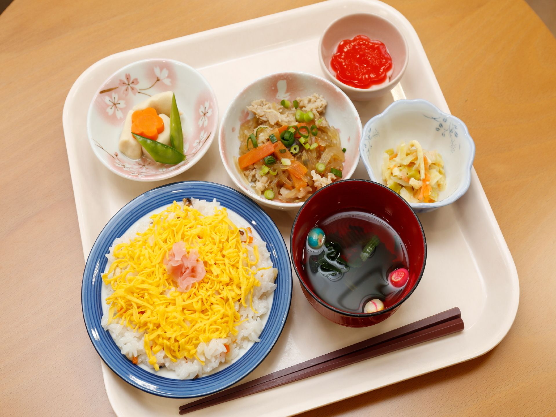 ある日の食事のメニュー。一人分がお盆にのっている。ちらし寿司を中心に、お吸い物や煮物、ゼリーなども一緒になっている。