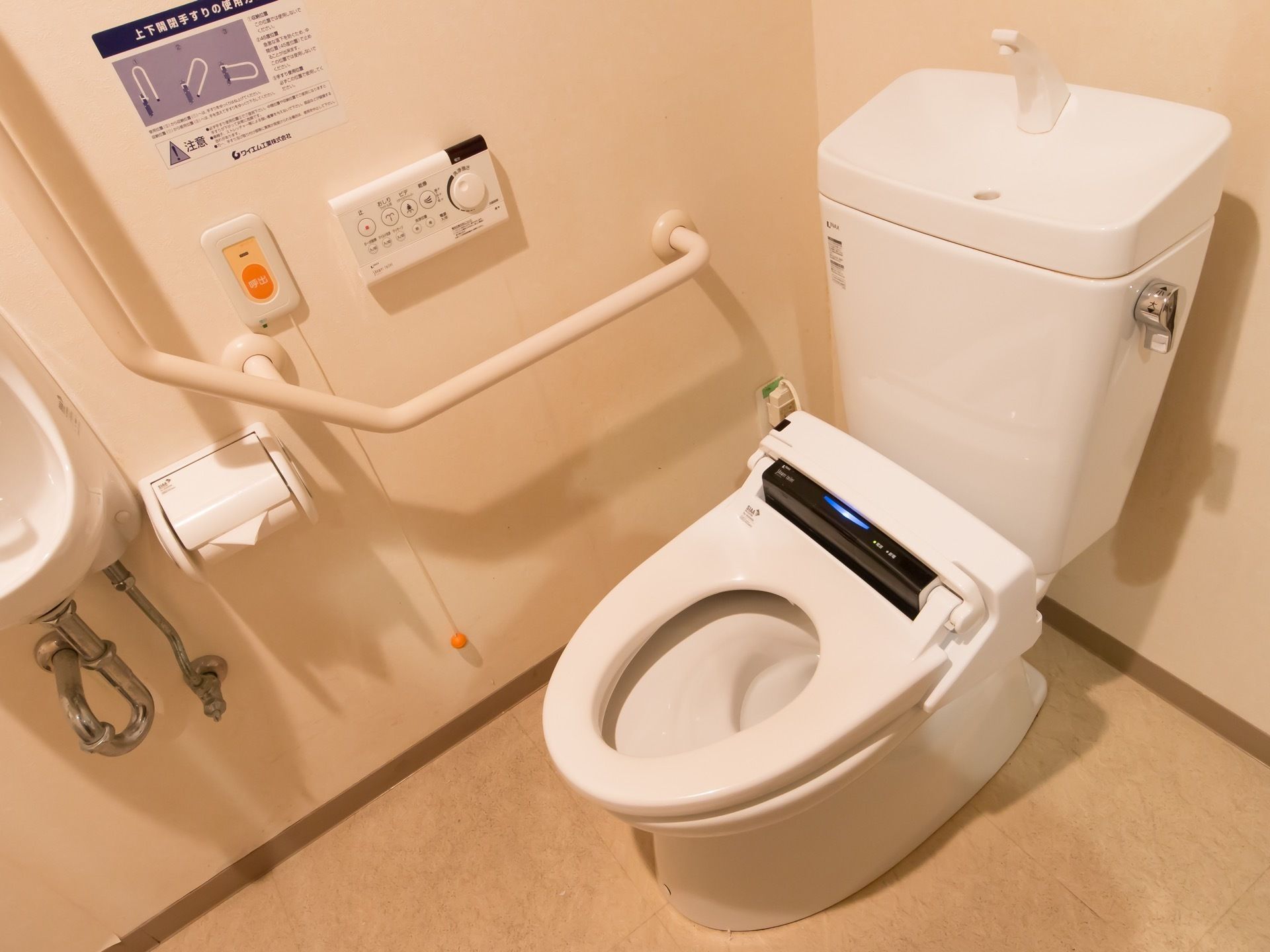 壁に緊急時呼出しボタンや手すりが備わったトイレである。可動式の手すりや簡易手洗い場が設置されている。