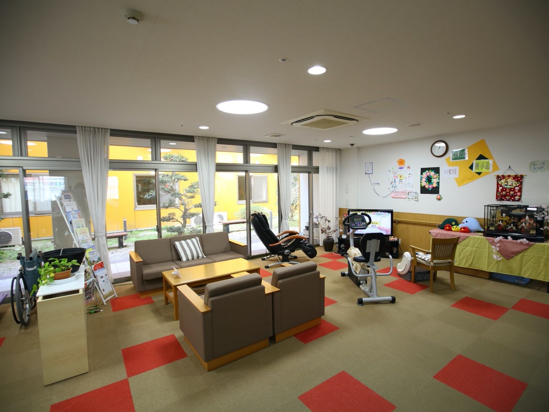 オレンジと緑のカーペットが敷かれている。テーブルとソファが設置されていて、奥にはテレビやトレーニングマシーンが置かれている。