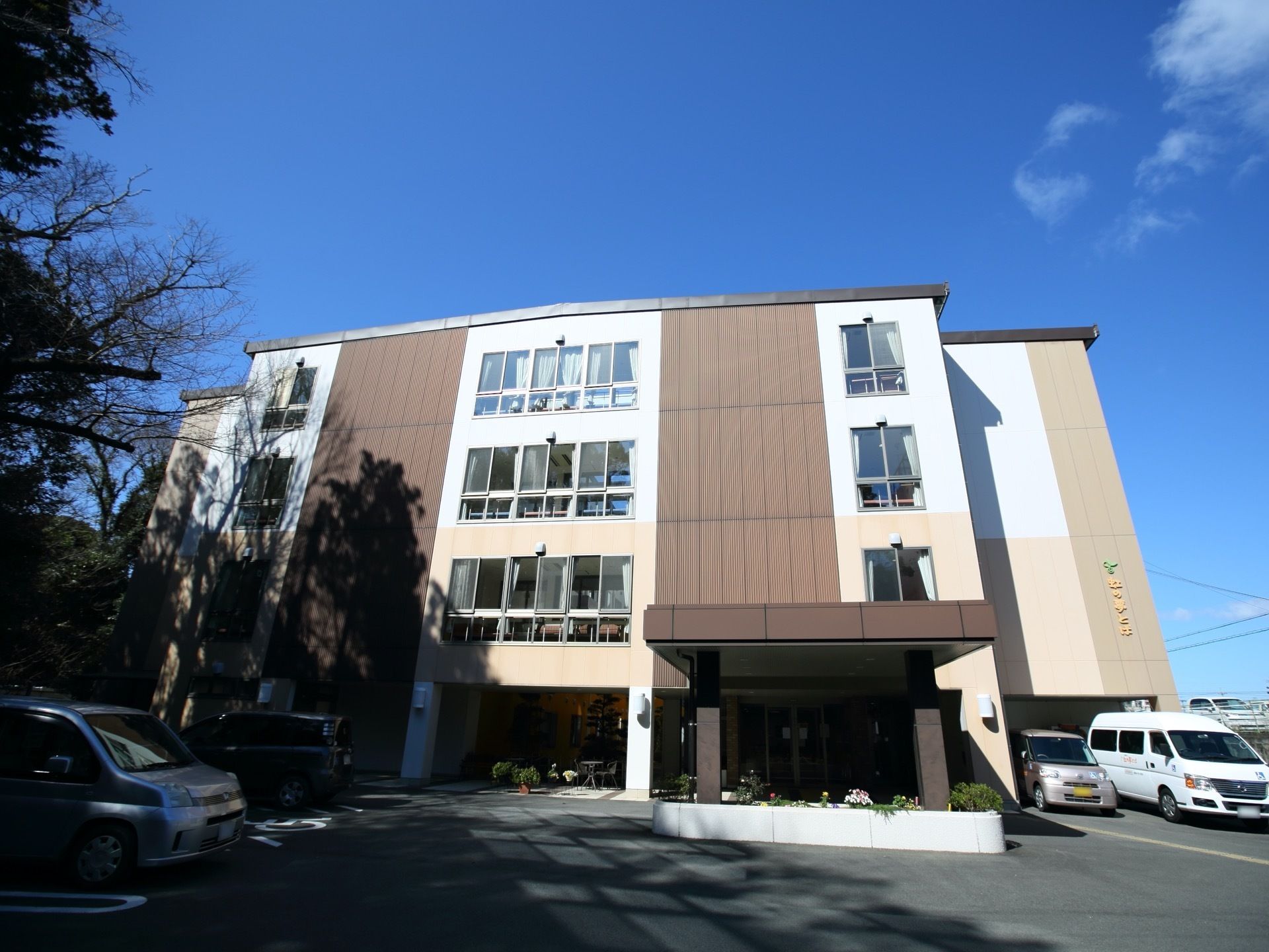 ブラウンを基調とした4階建ての建物である。建物の横には駐車スペースがあり、車が停められている。また、入口の前に花壇がある。