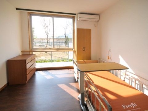 モダンな収納家具や、外観と揃いのオレンジのマットの介護用ベッドが置かれた居室。床から天井まである大きな窓の前には、手すりがつけられている。