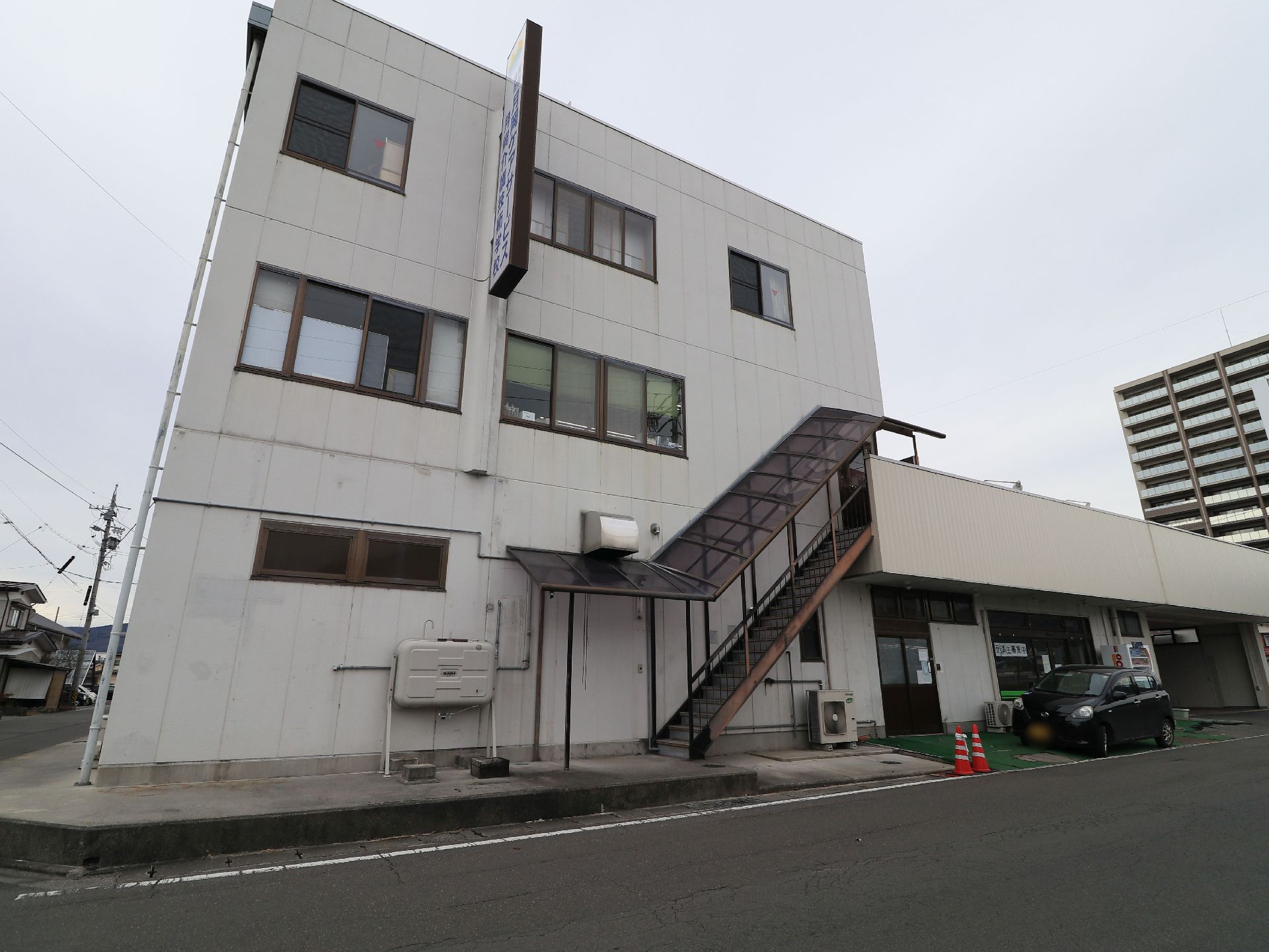 非常階段が設置されている建物である。施設の前には黒い車が１台停められており、自動販売機が置かれている。