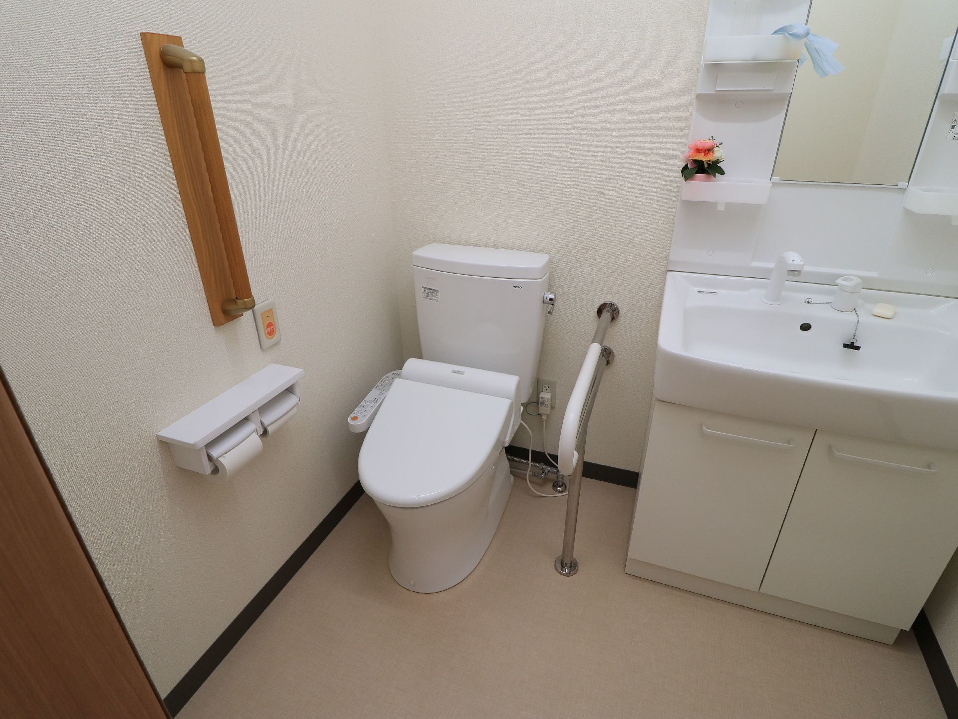 洗面台が備えつけられた個室トイレ。温水洗浄機能付きの便座で、壁には手すりやナースコールが設置されている。
