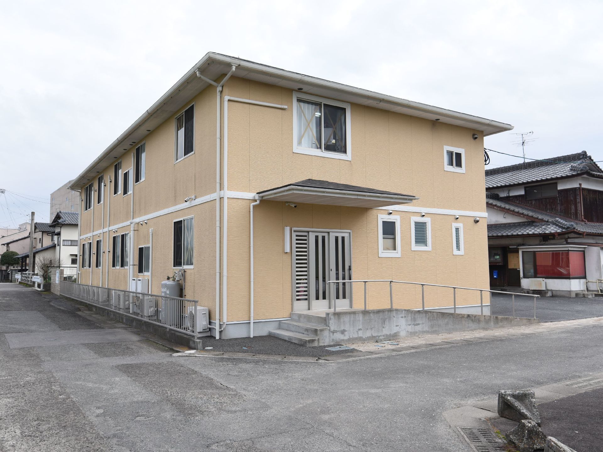 ２階建てで、クリーム色の優しい色合いのグループホーム。屋根付きの玄関で、横の外壁には照明が設置されている。