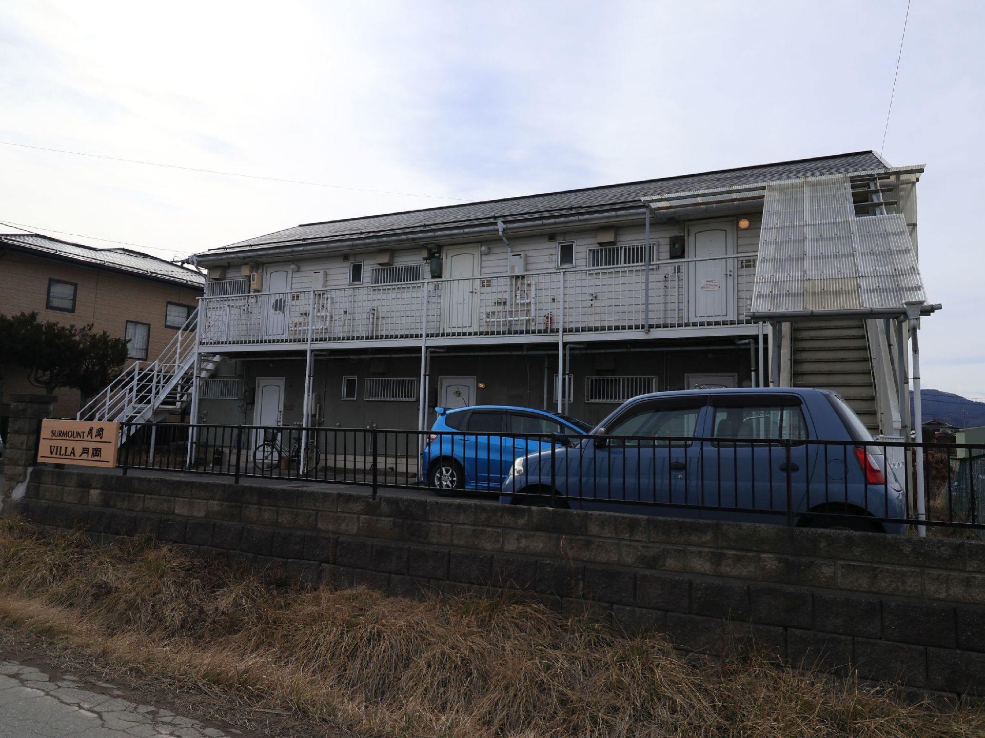 ２階建ての施設の建物が正面にあり、両側に外階段が設置されている。手前のフェンスの左手に施設名の入ったプレートが取りつけられている。