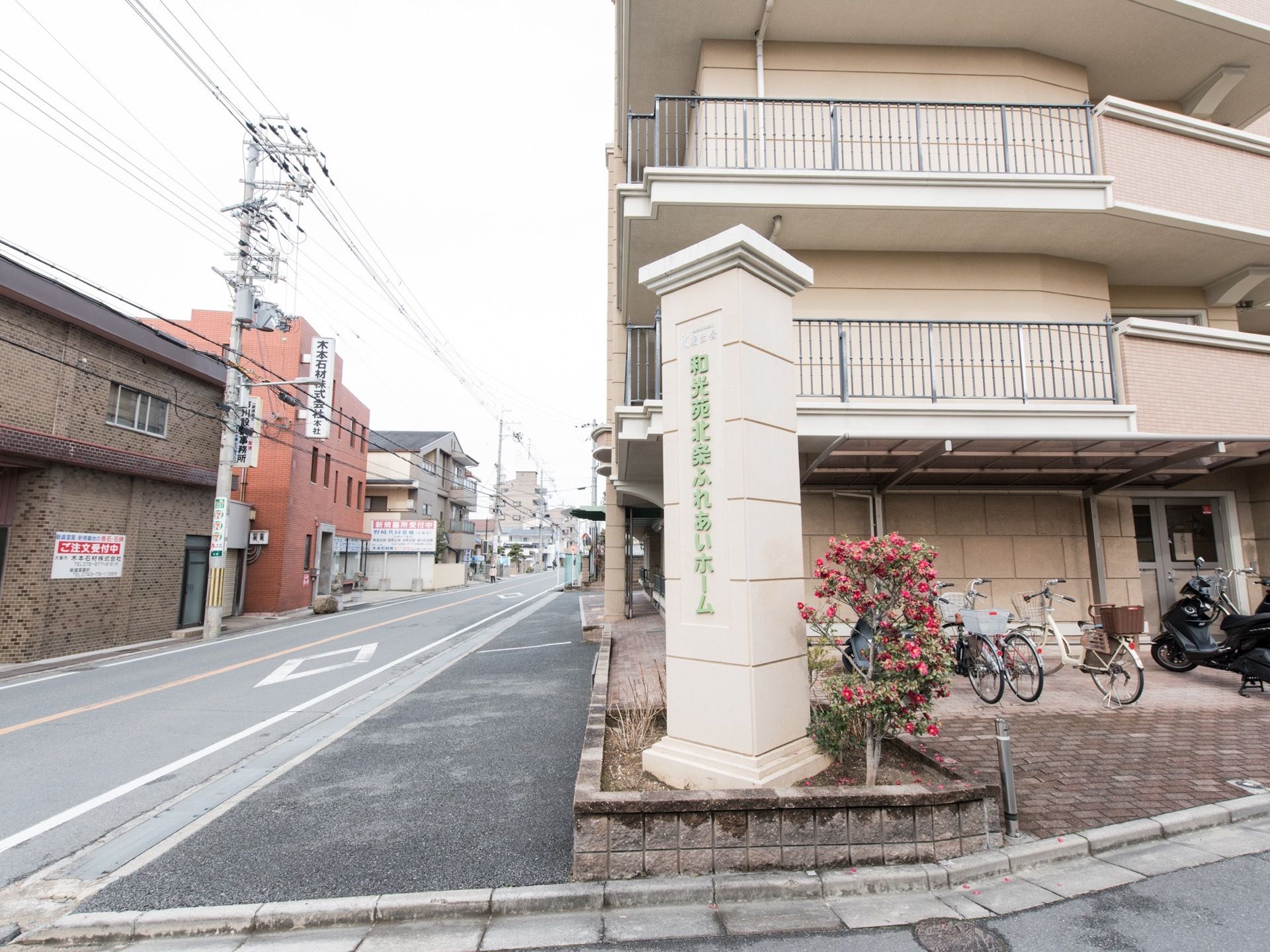 施設名称を表記した、石の柱が設置されている。近くに花が植えられている。建物の脇には、駐輪所がある。屋根が付いている。