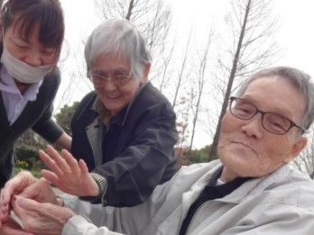 グレーの上着を着た高齢男性の手に、左側の高齢男性が手を置こうとしているのを介助するスタッフ