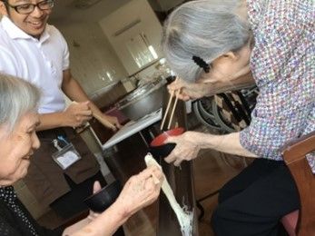 左にいる高齢女性が箸でもちをつまみ上げているのをお椀に取ろうとしている右側の高齢女性