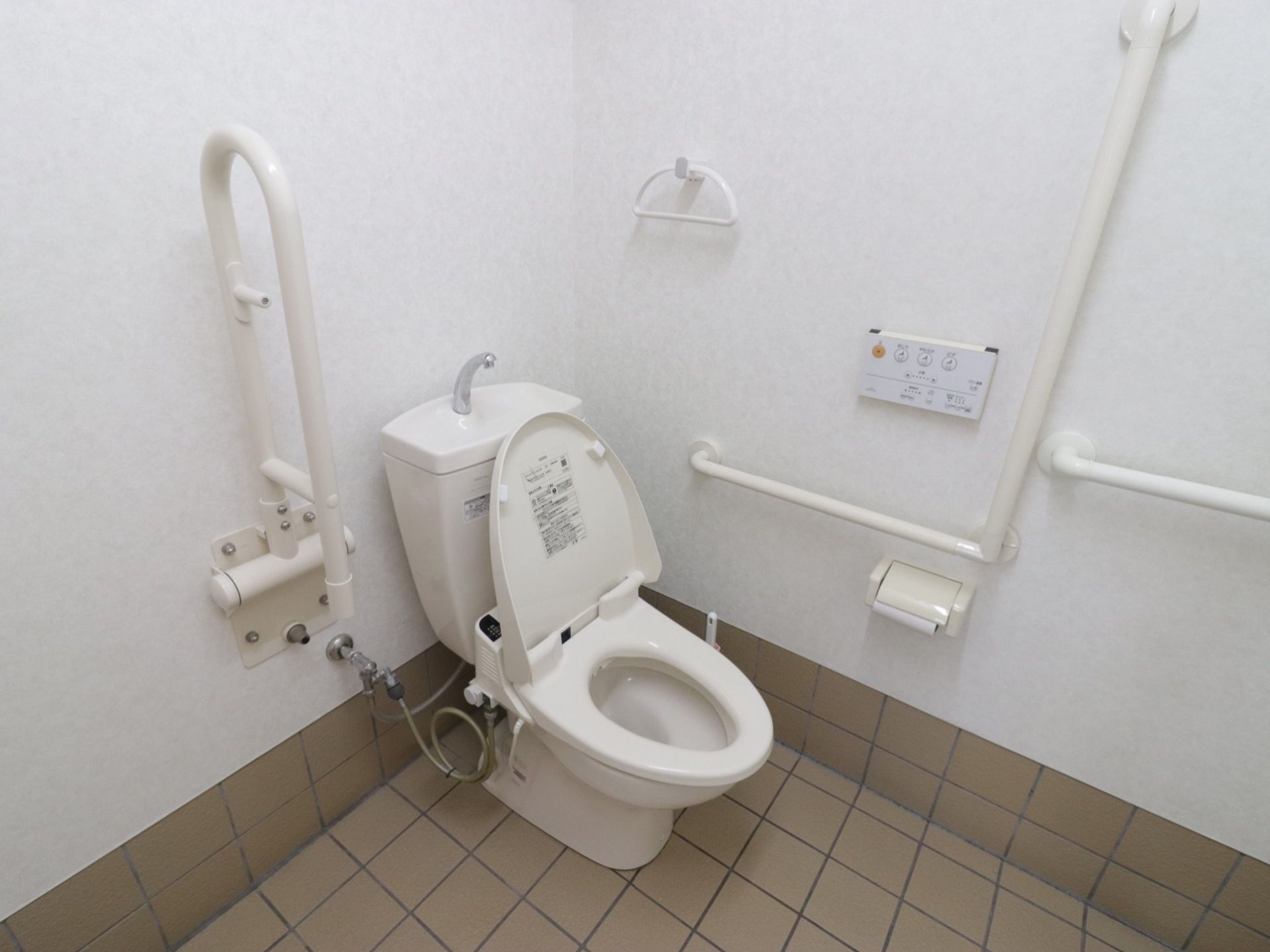 温水洗浄便座が設置されている。便座の両側に手すりがついている。片方はＬ字型である。タオルハンガーが壁についている。