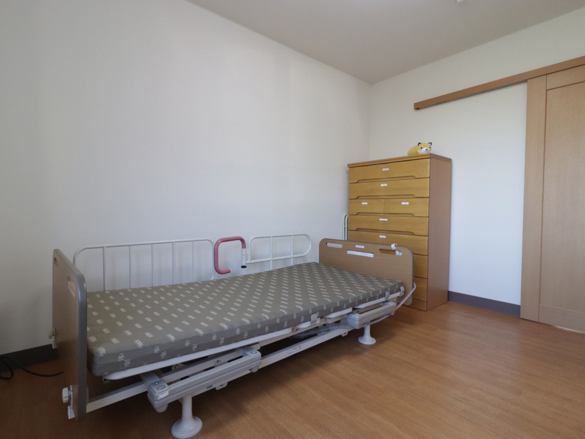 居室の壁は白色で、床はフローリングになっている。介護用の電動ベッド、高さのあるチェストが完備されている。