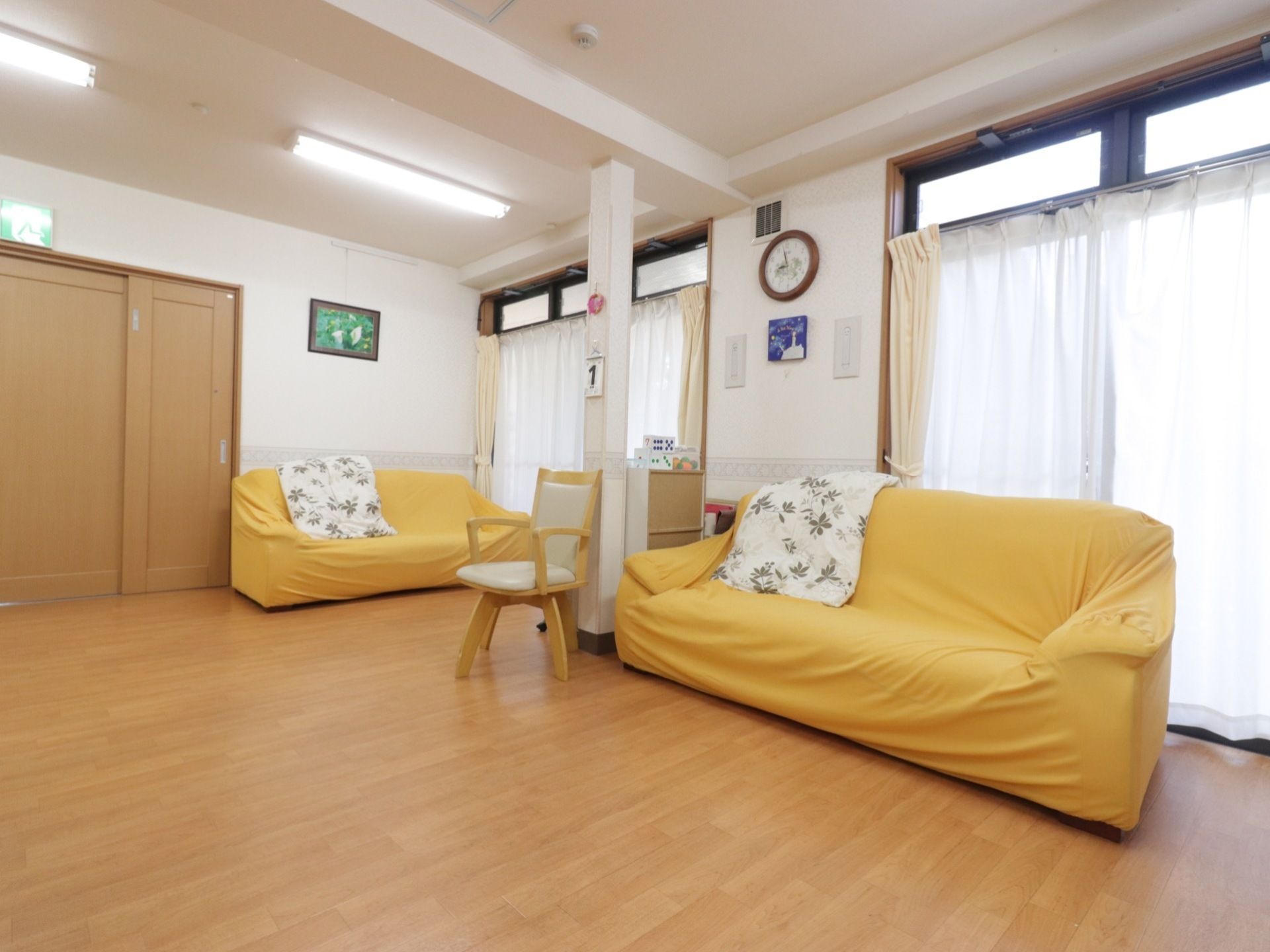 部屋の窓際にソファーが２つ、椅子が１脚置いてある。ソファーには黄色いカバーがかけられている。壁に絵が飾られている。