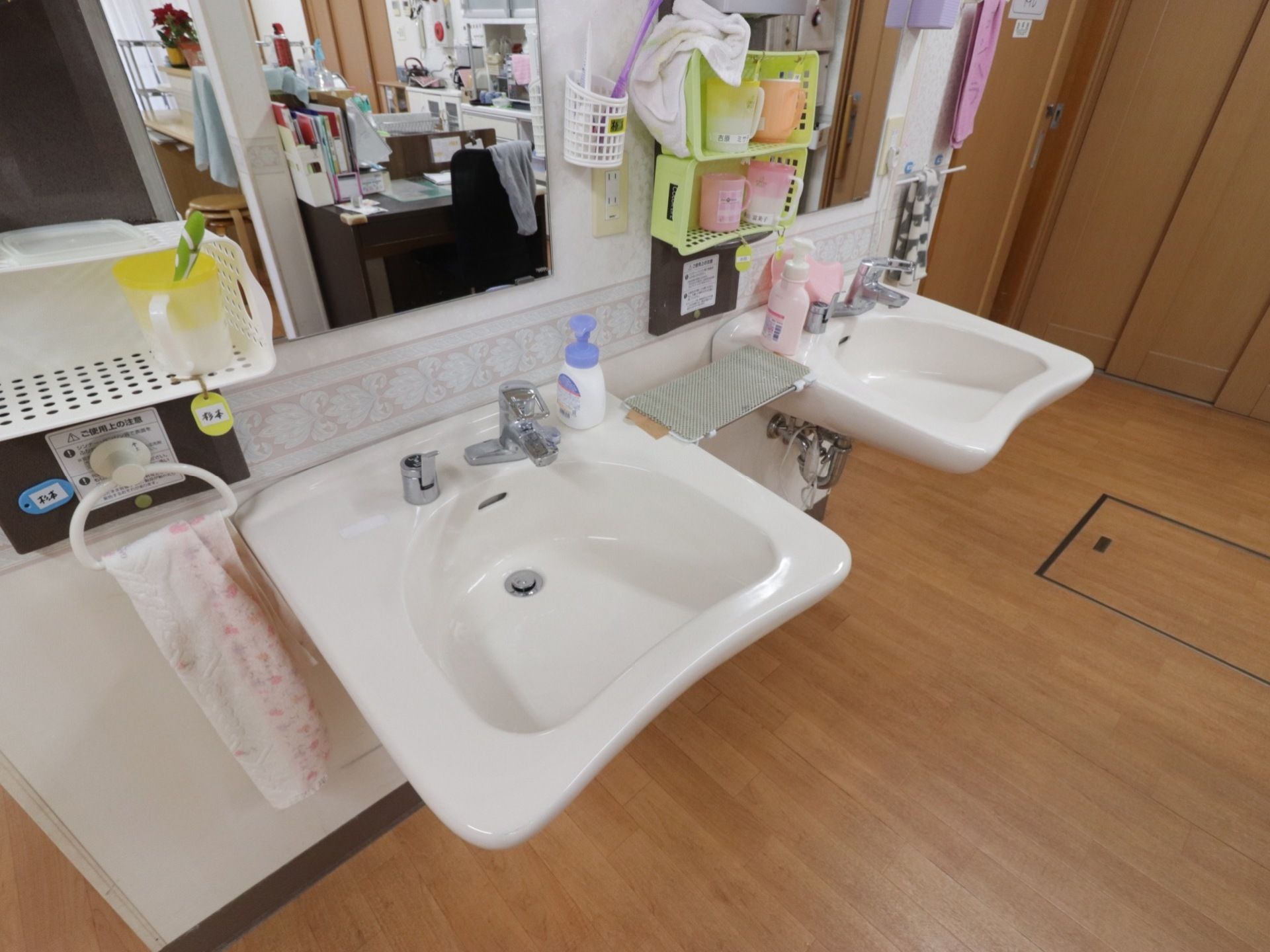 洗面台が２つ並んでいる。鏡があり、その前に入居者様の名前が書かれたコップと歯ブラシが置いてある。ハンドソープがそれぞれ置かれている。
