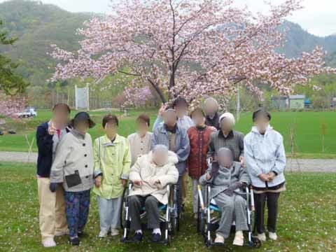 桜の気をバックに車椅子に座る高齢者2人を囲んで立つ10人の人物