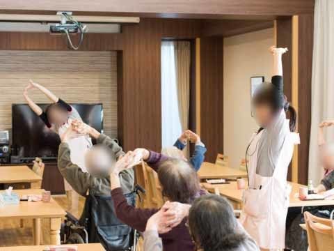 椅子に座って両手を上に伸ばす運動をする高齢者と一緒に運動しているスタッフ