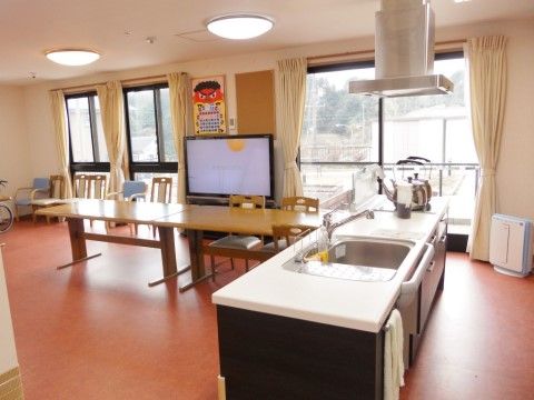 キッチンのあるリビングルームである。テーブル、椅子、テレビが置かれている。大きな窓と照明でとても明るい。