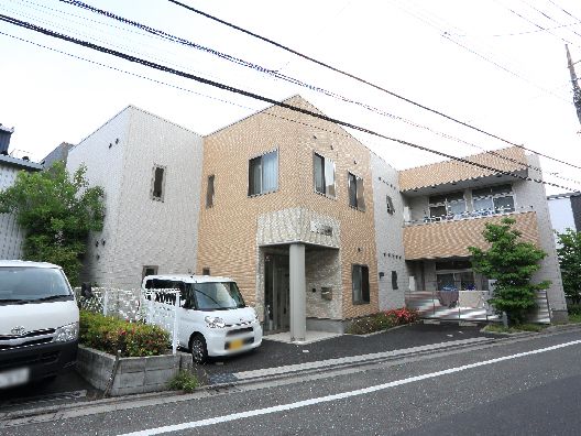 ２階建ての建物はベージュ系の外壁で敷地内には駐車場があり、車が止まっている。