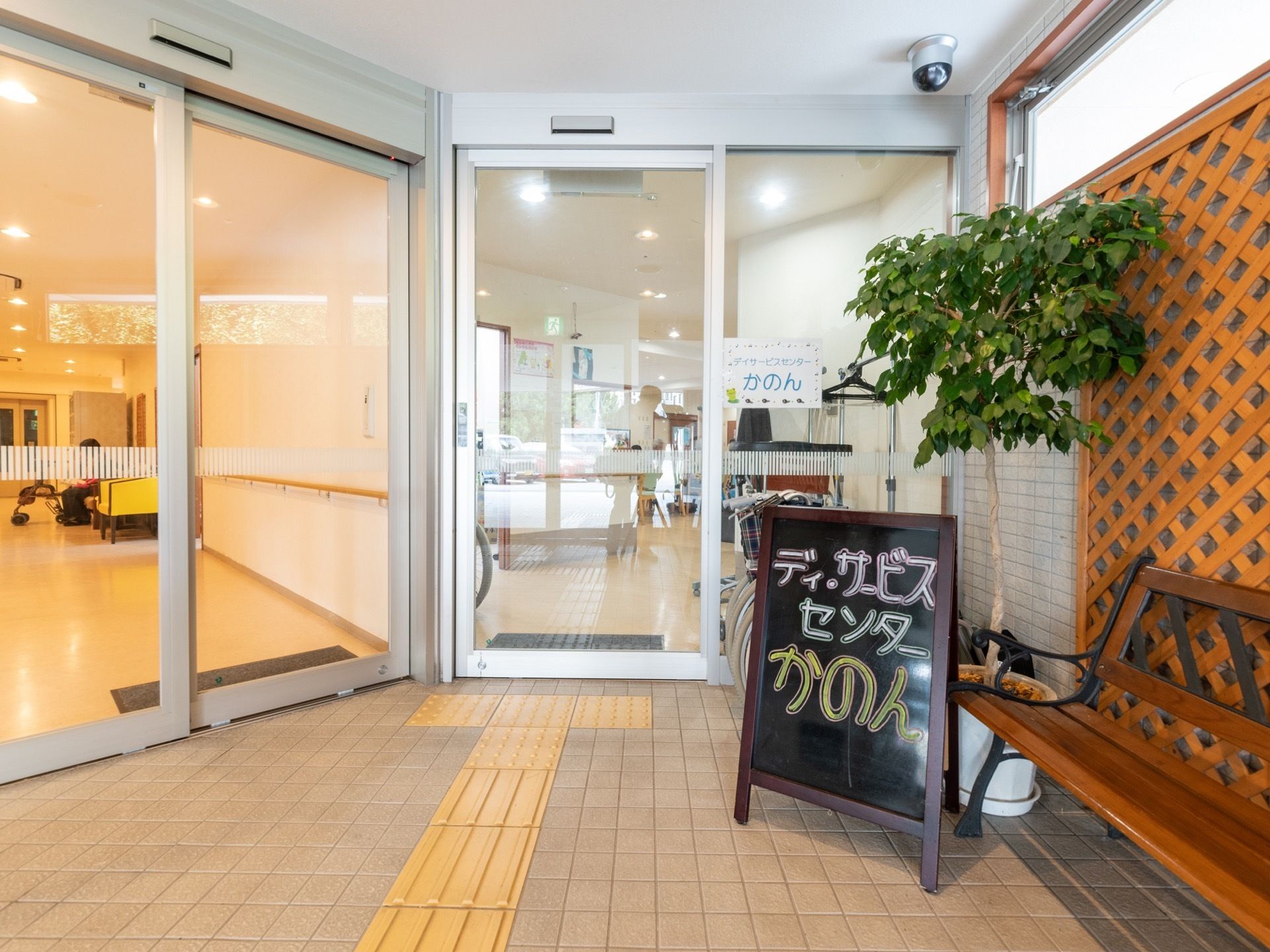 ガラスの自動ドアが二つ並んでいる。右側の扉の前にはデイサービスと書かれたブラックボードが置かれている。