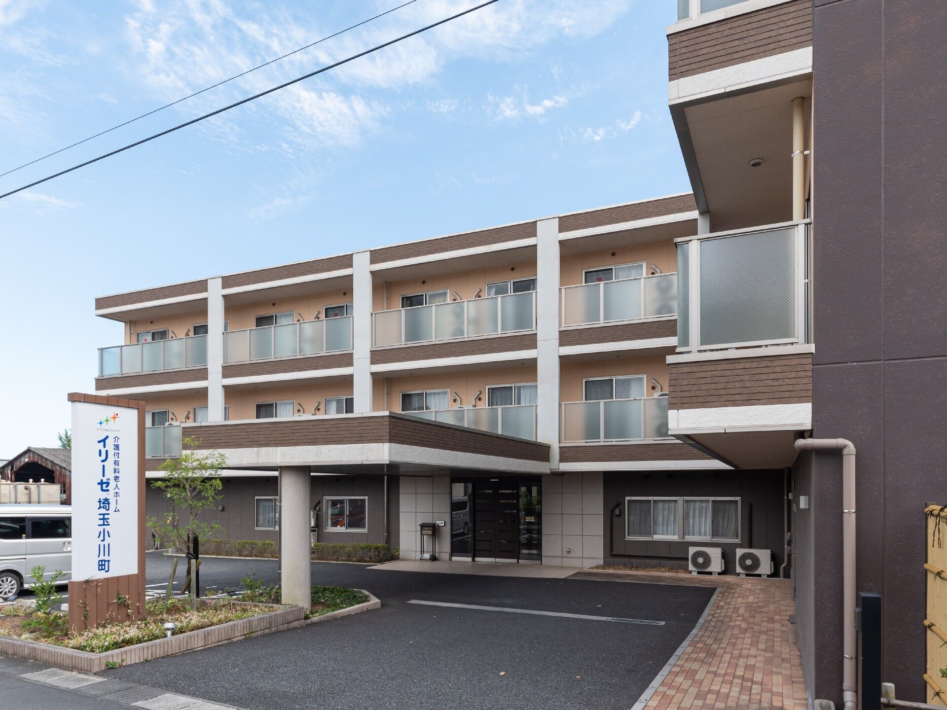 白にダークブラウンの縦のラインがモダンな施設の外観。エントランス前には屋根が設置され、車から濡れずに施設に入ることができる。